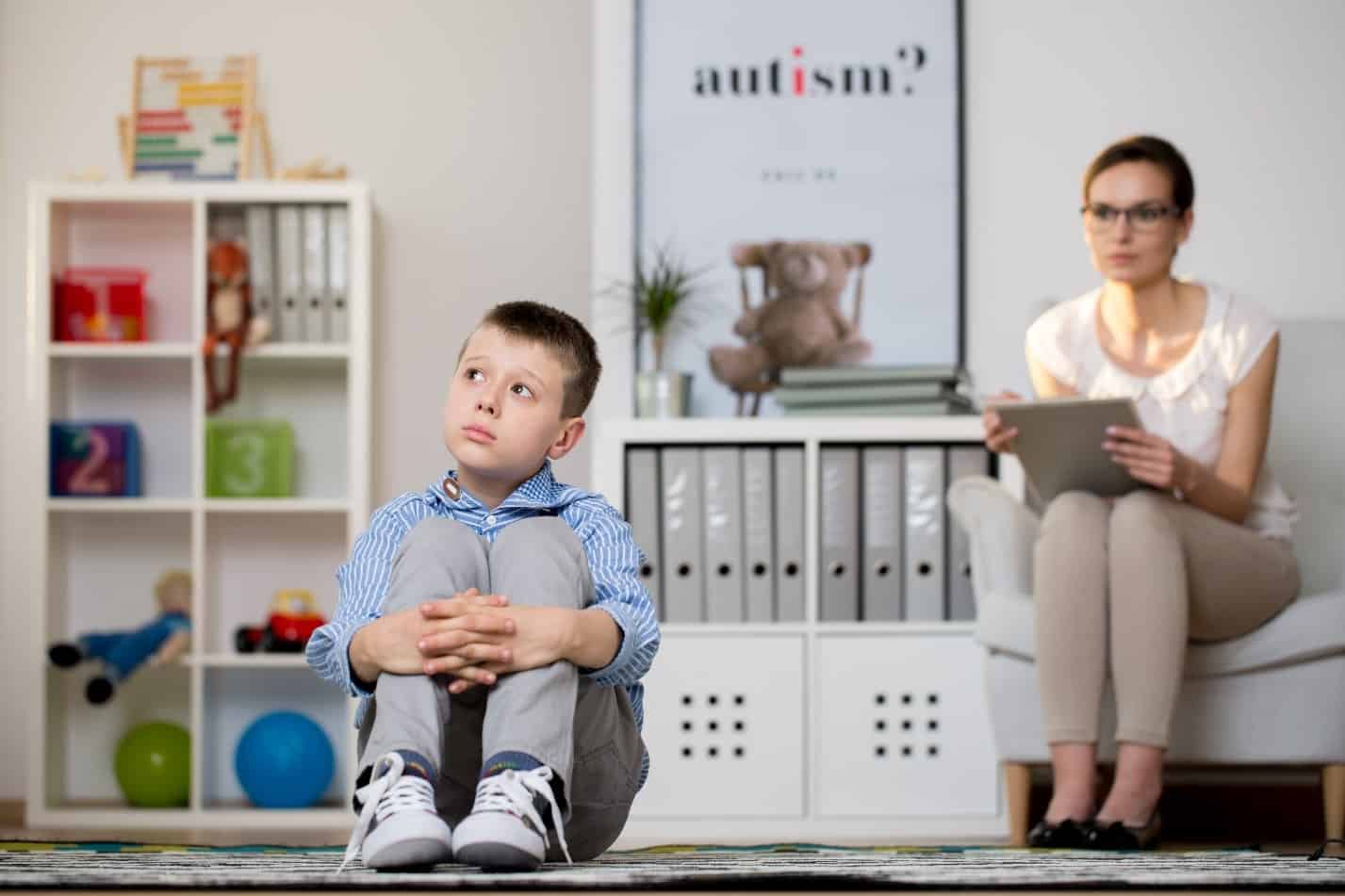 Child and therapist seated in in front of bookshelves and a poster featuring the word autism with a question mark.