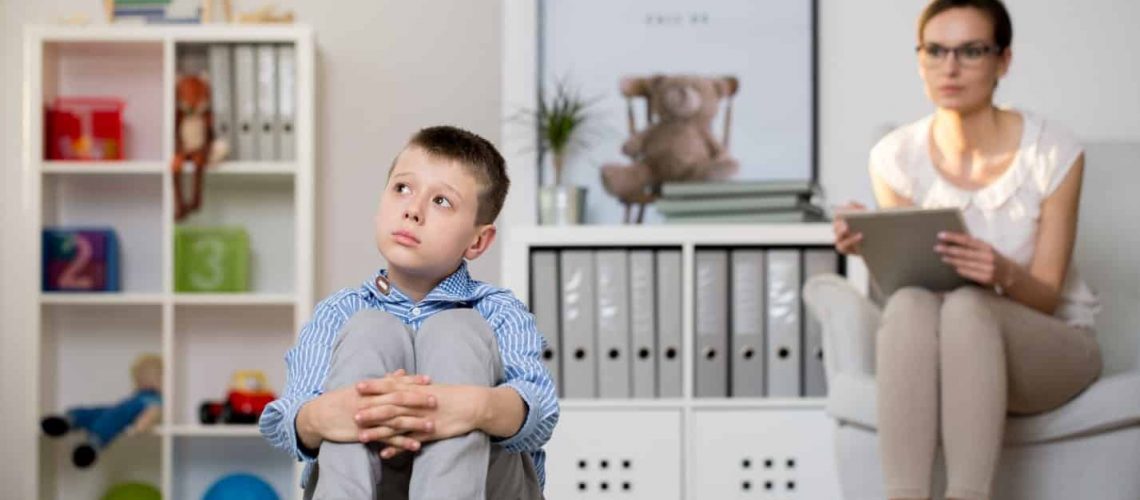 Child and therapist seated in in front of bookshelves and a poster featuring the word autism with a question mark.